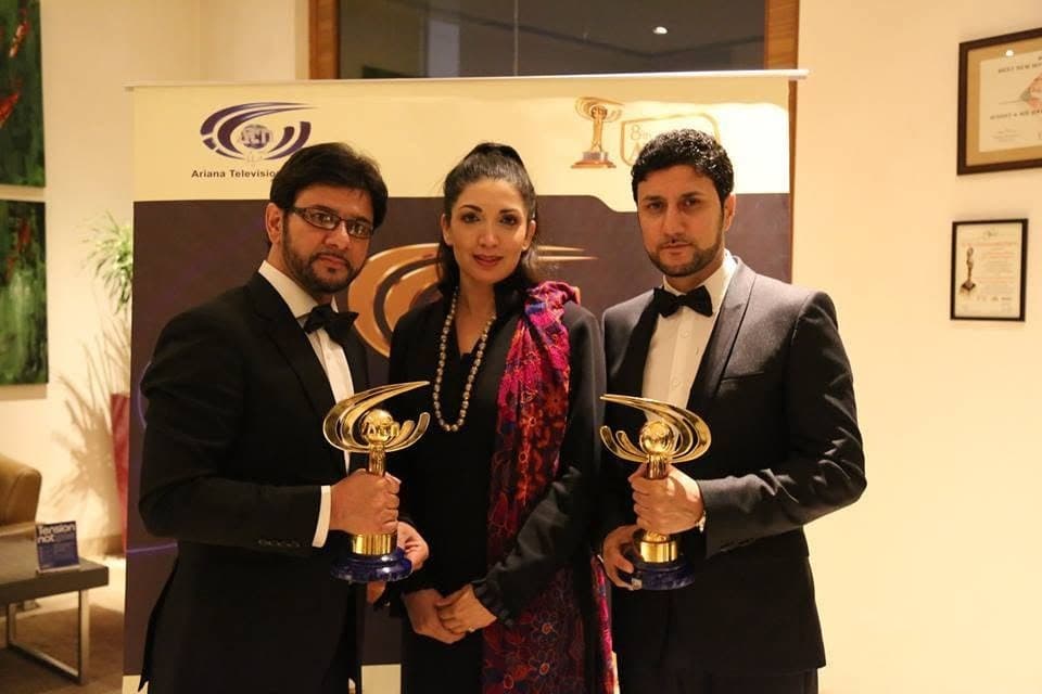 Taher Shubab in a tuxedo holding an award trophy at the Ariana Television awards, beside two guests