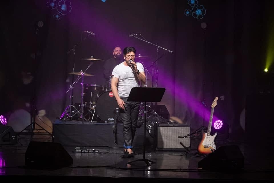 Taher Shubab singing on a concert stage bathed in purple light, with a drummer and an electric guitar behind him