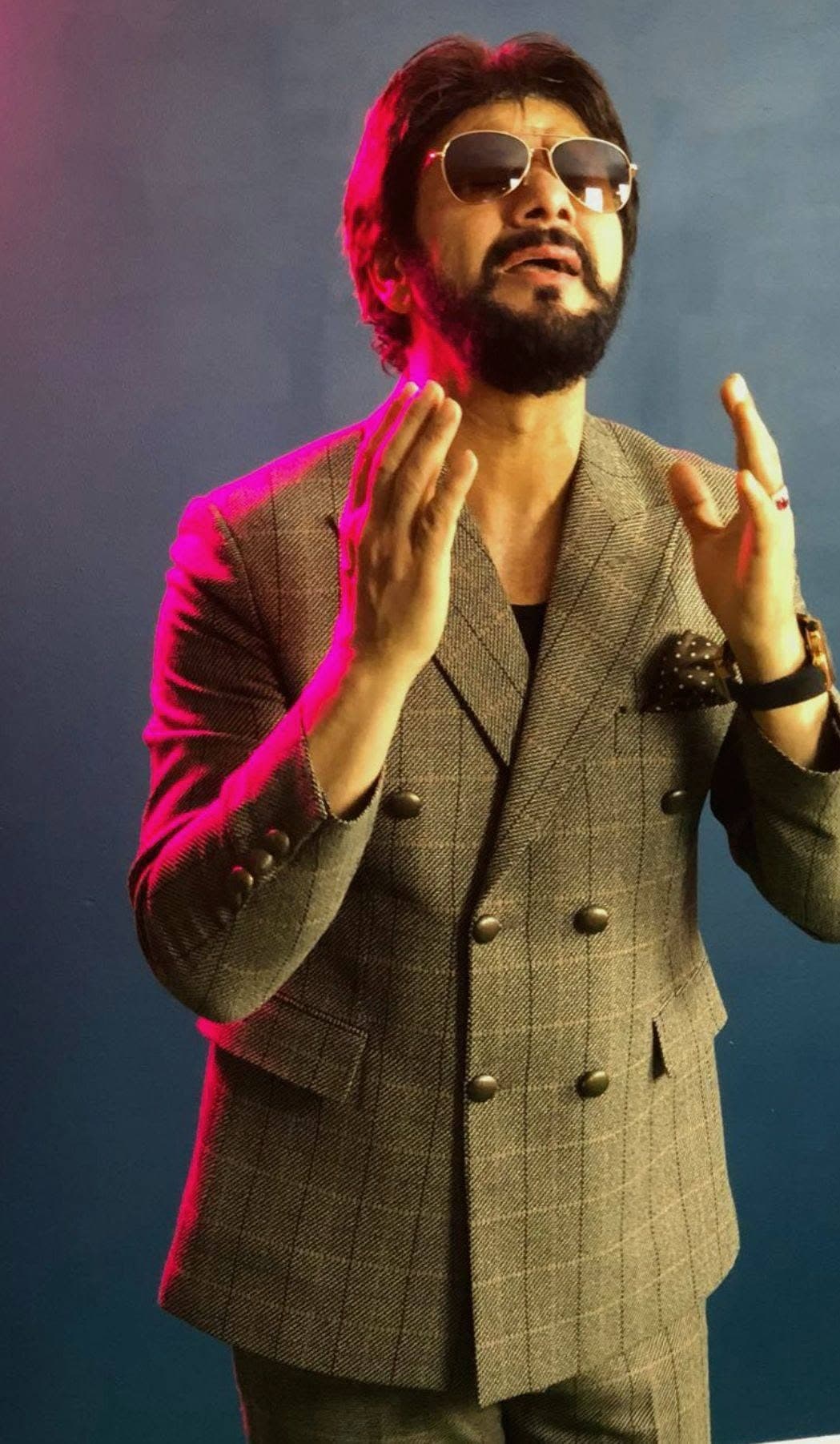 Studio portrait of Taher Shubab singing in a grey double-breasted suit and aviator sunglasses under pink light