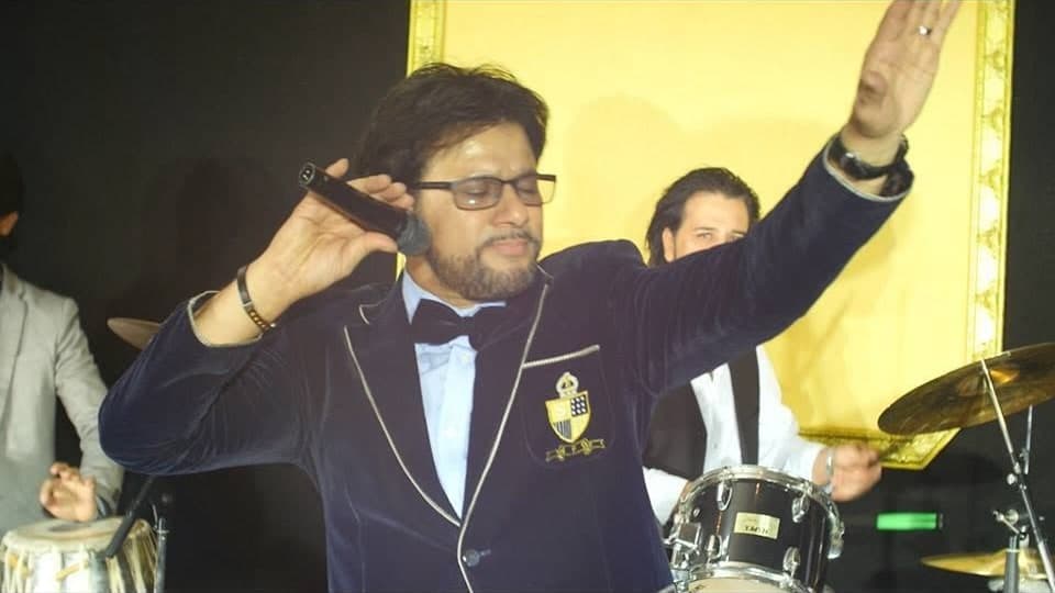 Taher Shubab in a navy velvet blazer and bow tie raising his hand while singing with his band